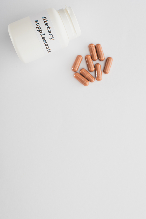 Top view of container with dietary supplements lettering and brown capsules on white background