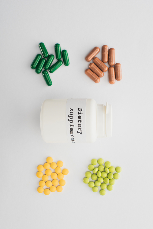 Top view of container with dietary supplements lettering with pills and capsules on white background