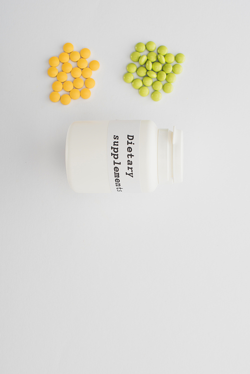 Top view of container with dietary supplements lettering and colorful pills on white background
