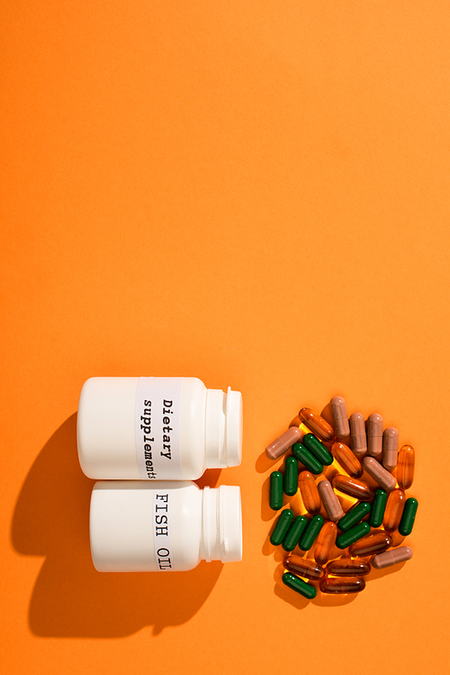 Top view of containers with fish oil and dietary supplements lettering near colorful capsules on orange background