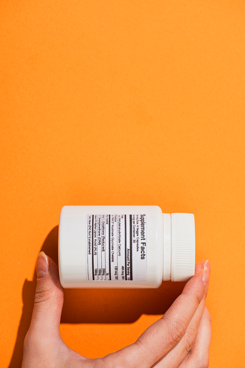 Partial view of woman holding white container with dietary supplements on orange background