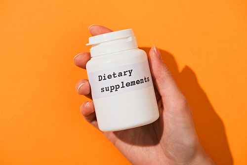Partial view of woman holding container with dietary supplements lettering on orange background