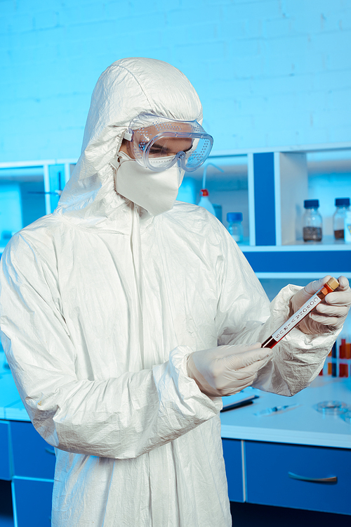 scientist in hazmat suit and goggles holding test tube with coronavirus lettering