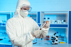 scientist in hazmat suit, latex gloves and goggles holding test tube with coronavirus lettering