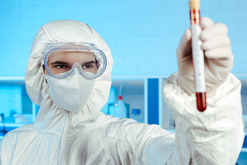 selective focus of scientist in hazmat suit and goggles holding test tube with coronavirus lettering