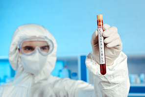 selective focus of scientist in hazmat suit, medical mask and goggles holding test tube with coronavirus lettering
