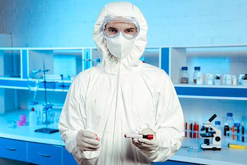 scientist in hazmat suit, medical mask and goggles holding sample with coronavirus lettering