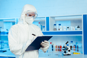 scientist in hazmat suit and goggles writing while holding clipboard near microscope