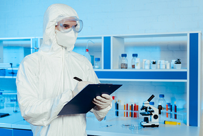 scientist in hazmat suit and goggles holding clipboard and pen near microscope
