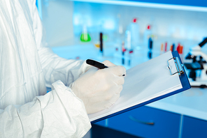 cropped view of scientist in hazmat suit writing on blank paper while holding clipboard