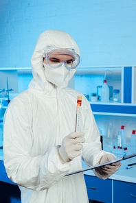 scientist in hazmat suit and goggles holding test tube with coronavirus lettering and clipboard