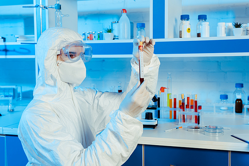 scientist in hazmat suit and latex gloves holding test tube with coronavirus lettering