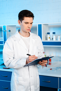 handsome scientist in white coat holding clipboard and pen in laboratory