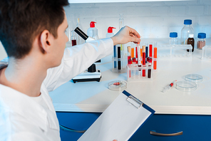 selective focus of scientist in white coat holding clipboard and touching test tube