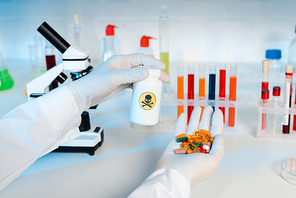 cropped view of scientist in latex gloves holding bottle with danger sign and pills