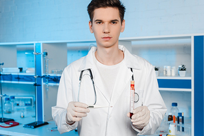 handsome scientist in latex gloves holding digital thermometer and test tube with coronavirus lettering
