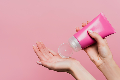 cropped view of woman holding tube while applying lotion on hand isolated on pink