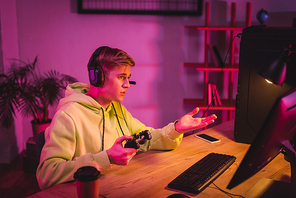 KYIV, UKRAINE - AUGUST 21, 2020: Gamer in headset holding joystick while pointing at computer monitor near smartphone and coffee to go on blurred foreground