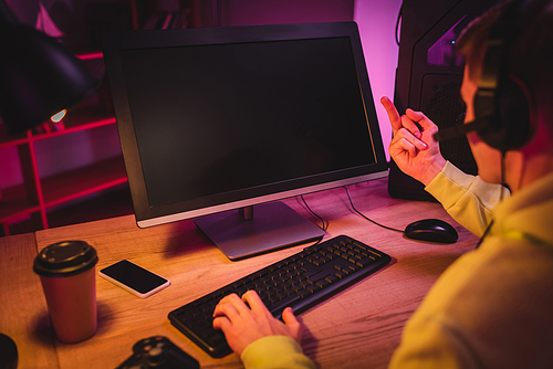 Player in headset showing middle finger at computer monitor while playing video game near smartphone and takeaway coffee on blurred foreground