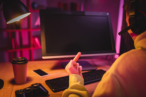 KYIV, UKRAINE - AUGUST 21, 2020: Gamer in headset showing middle finger near joystick, coffee to go and computer on blurred background