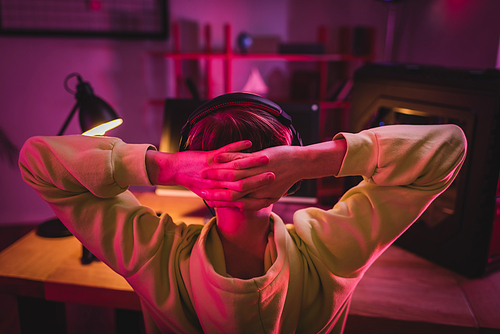 Back view of gamer in headset with hands near head sitting near computer on blurred background