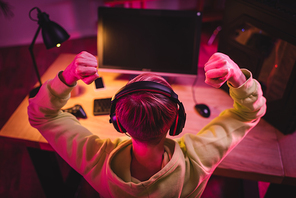 Overhead view of gamer in headphones showing yeah gesture near computer and joystick on blurred background