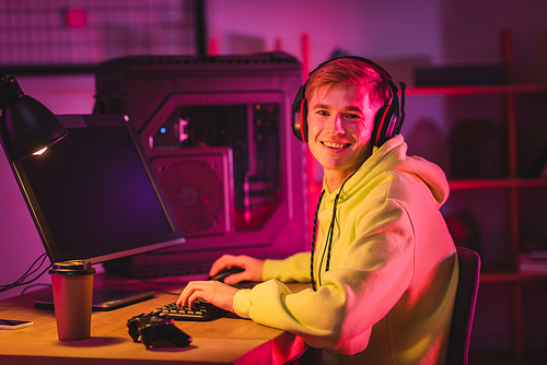KYIV, UKRAINE - AUGUST 21, 2020: Side view of smiling gamer in headphones using computer keyboard near joystick and coffee to go on blurred foreground