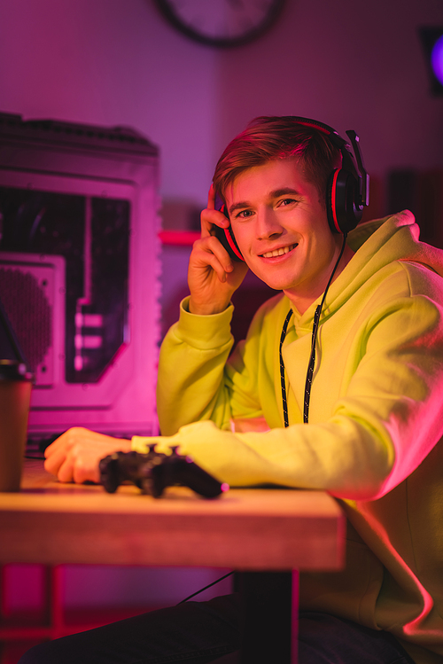 Smiling gamer holding headphones near joystick and coffee to go on blurred foreground