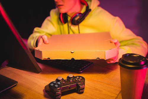 KYIV, UKRAINE - AUGUST 21, 2020: Joystick and coffee to go near gamer in headphones holding pizza box near computer on blurred background