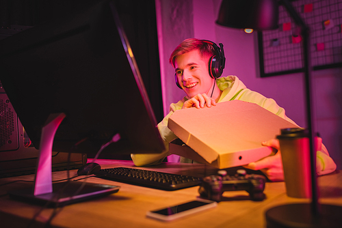 Smiling gamer in headphones holding pizza box near computer, joystick and smartphone on blurred foreground