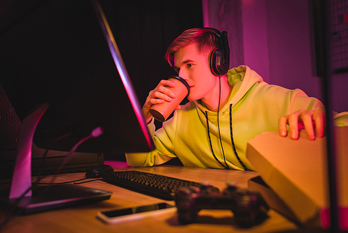 Young gamer in headphones drinking coffee to go and holding pizza box near computer and joystick on blurred foreground