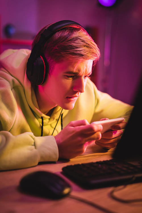 Gamer in headphones playing game on smartphone near computer on blurred foreground
