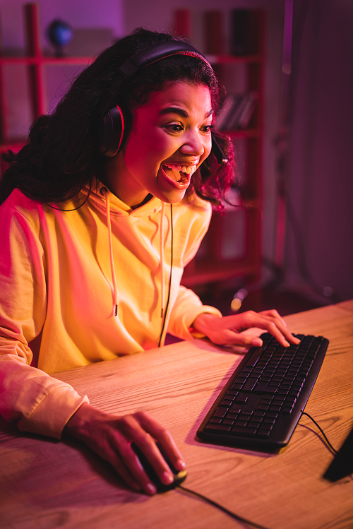 Excited african american gamer in headset using computer keyboard and mouse on blurred foreground