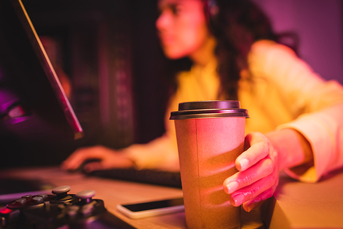 KYIV, UKRAINE - AUGUST 21, 2020: Coffee to go near joystick and gamer using computer on blurred background
