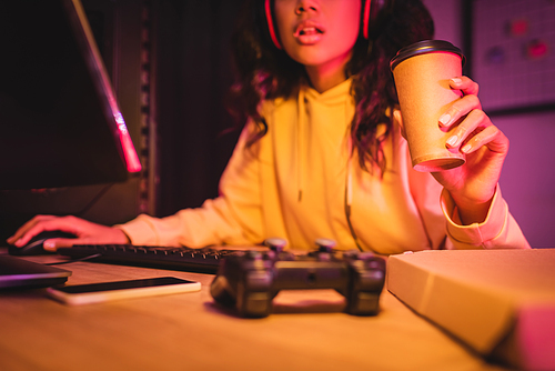 Cropped view of takeaway coffee in hand of african american gamer using computer near joystick on table