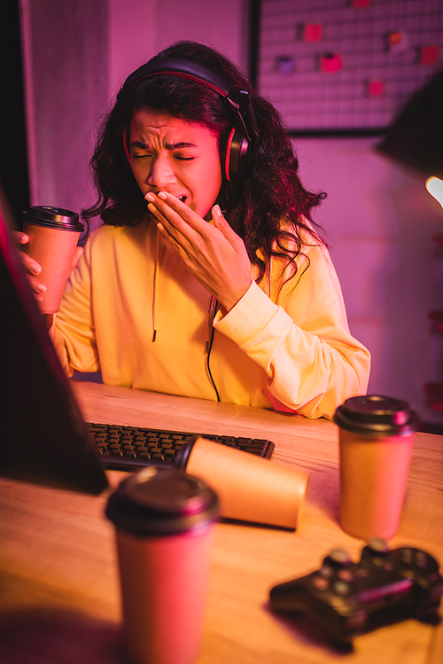 Tired african american gamer yawning while holding coffee to go near computer and joystick on blurred foreground