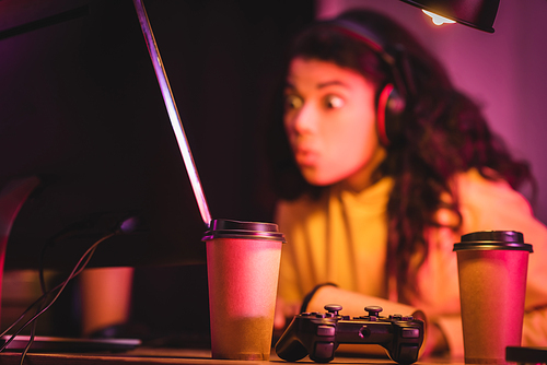 KYIV, UKRAINE - AUGUST 21, 2020: Joystick and coffee to go near excited african american gamer and computer on blurred background