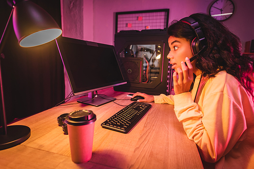 Excited african american gamer in headset using computer with blank screen near lamp and coffee to go on blurred foreground