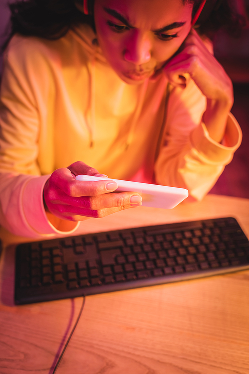 Pensive african american gamer using smartphone near computer keyboard on blurred background