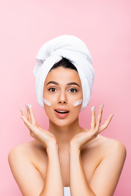 surprised beautiful woman with towel on hair and cosmetic cream on face isolated on pink
