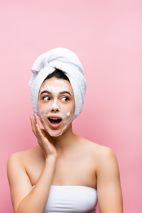 shocked beautiful woman with towel on hair and foam on face isolated on pink