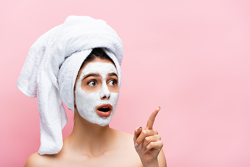 shocked beautiful woman with towel on head and clay mask on face pointing aside isolated on pink