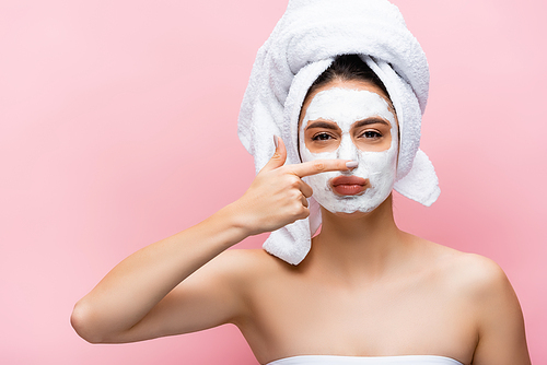 beautiful woman with towel on head and clay mask on face touching nose isolated on pink