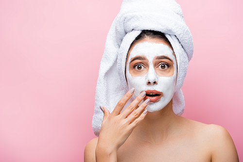 shocked beautiful woman with towel on head and clay mask on face isolated on pink