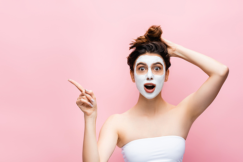 shocked beautiful woman with clay mask on face pointing aside isolated on pink