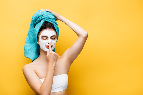 beautiful woman with blue towel on hair and clay mask on face touching nose isolated on yellow