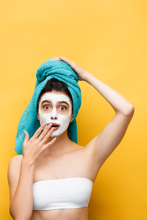 shocked beautiful woman with blue towel on hair and clay mask on face isolated on yellow