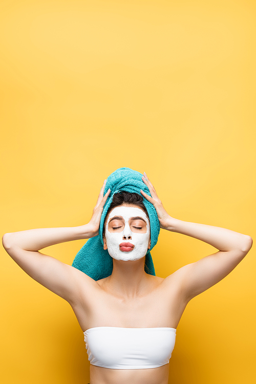 beautiful woman with blue towel on hair and clay mask on face blowing kiss isolated on yellow