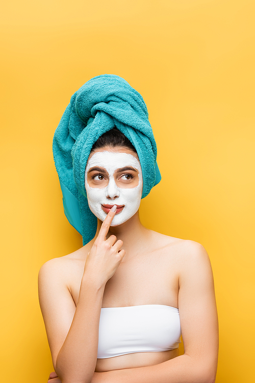 pensive beautiful woman with blue towel on hair and clay mask on face isolated on yellow