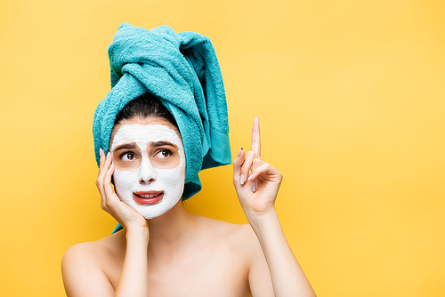 confused beautiful woman with blue towel on hair and clay mask on face pointing up isolated on yellow